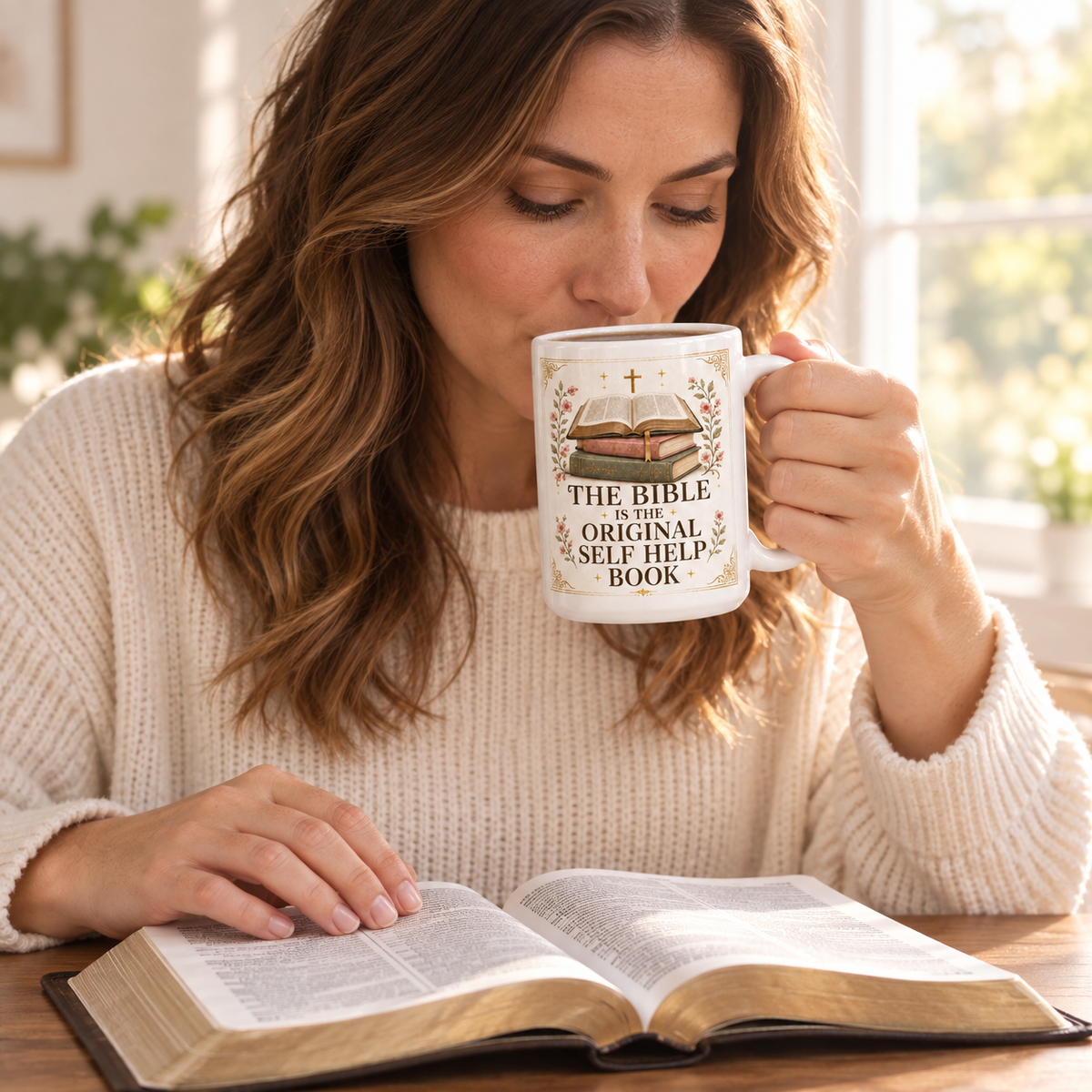A woman in a cream knit sweater reads in a bright room while holding a white ceramic mug with a graphic of a Bible and the text 'THE BIBLE IS THE ORIGINAL SELF HELP BOOK'. An open book lies on the table in front of her.